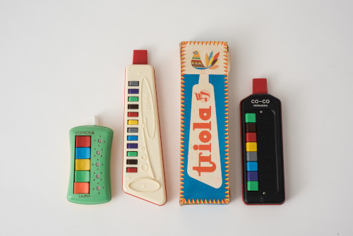 Foto: 3 nebeneinander angeordnete Blasinstrumente aus Plastik mit bunten Tasten und eine Verpackung mit der Aufschrift "Triola".